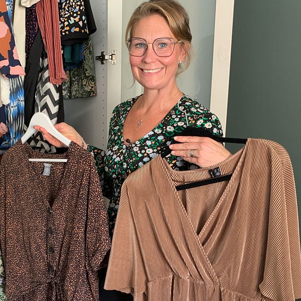 Anna Sanneviken som inte handlat nya kläder på flera år står framför en garderob och håller upp två klänningar, en mönstrad och en i beige, medan kläder hänger bakom henne. Garderoben är fylld med olika plagg, inklusive toppar och kjolar.
