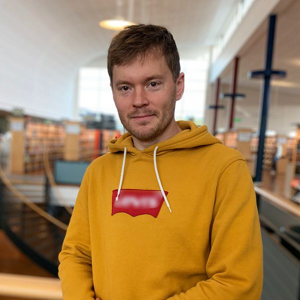 En man tittar rakt in i kameran. Han har en gul tröja och i bakgrunden är det ett bibliotek.