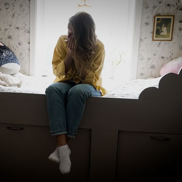 Ung person sitter ensam på en säng i ett dunkelt rum, ser nedstämd ut.