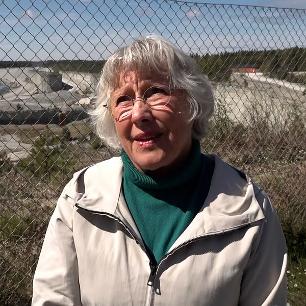 Person med vitt hår står utomhus vid stängsel, hav och industriområde syns i bakgrunden.