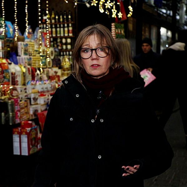 Person står framför en färgglad julmarknad med olika presenter och dekorationer i bakgrunden, belyst av julbelysning.
