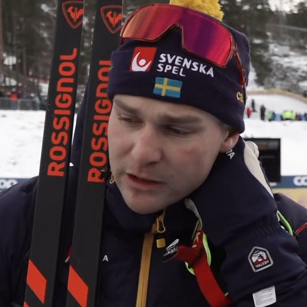 En skidåkare i tävlingsdräkt håller i sina skidor med Rossignol-logotyp. I bakgrunden syns en vintersportarena med snö och andra åkare.