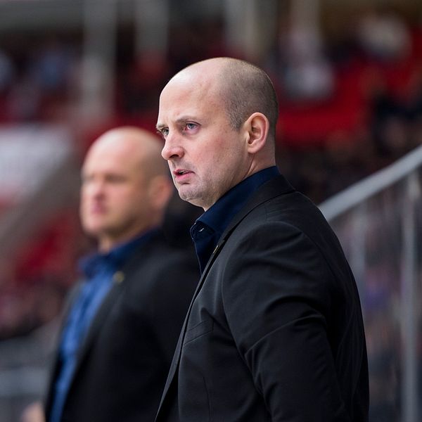 Två män står vid sidan av en hockeyrink, klädda i kostymer. I bakgrunden syns en publik i röda läktarsektioner, vilket skapar en sportig atmosfär. Den ena mannen ser ut att vara i fokus, medan den andra står lite längre tillbaka.