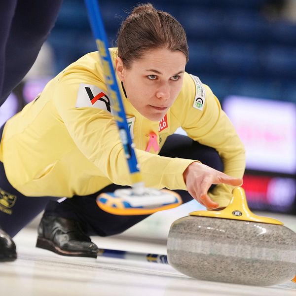 Anna Hasselborg i en gul tröja ligger på knä och siktar på en curlingsten som glider över isen. En annan spelare syns delvis i bakgrunden, klädd i mörka byxor. Hallen är inredd för en curlingmatch med oskarpa åskådare i bakgrunden.