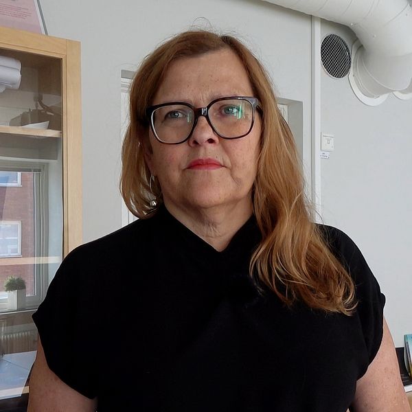 En kvinna med långt brunt utslaget hår som faller över vänster axel, hon har svarta glasögon och svart v-ringad tröja som går högt upp mot halsen.
