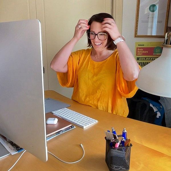 Person i gul tröja sitter vid skrivbord med dator, lyfter upp en surfplatta eller bok.