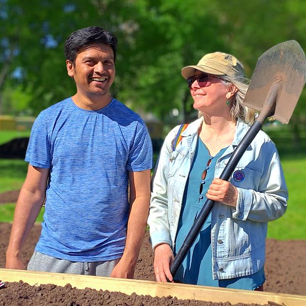 Två personer, en man med blå tröja som skrattar och en kvinna med spade, står framför odlingslåda
