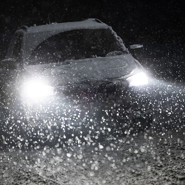 En bil kör med tända framljus i mörkret. Omkring bilen faller snön.