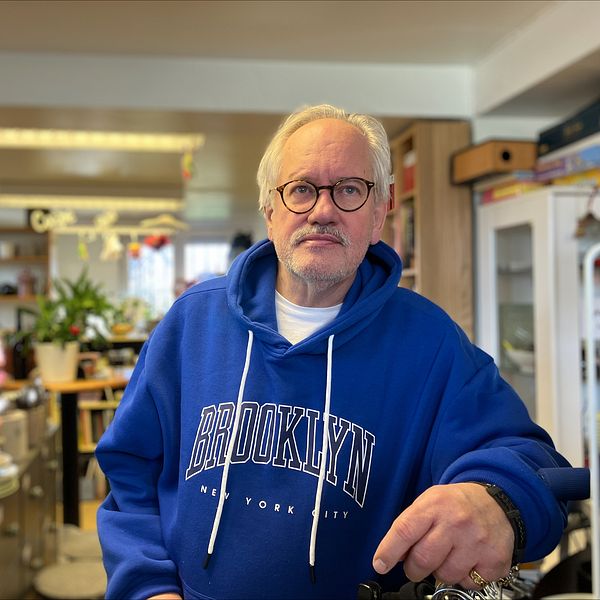 En äldre man i en blå Brooklyn-hoodie står i en återbrukslokal med uppställda tallrikar och inredning i bakgrunden.