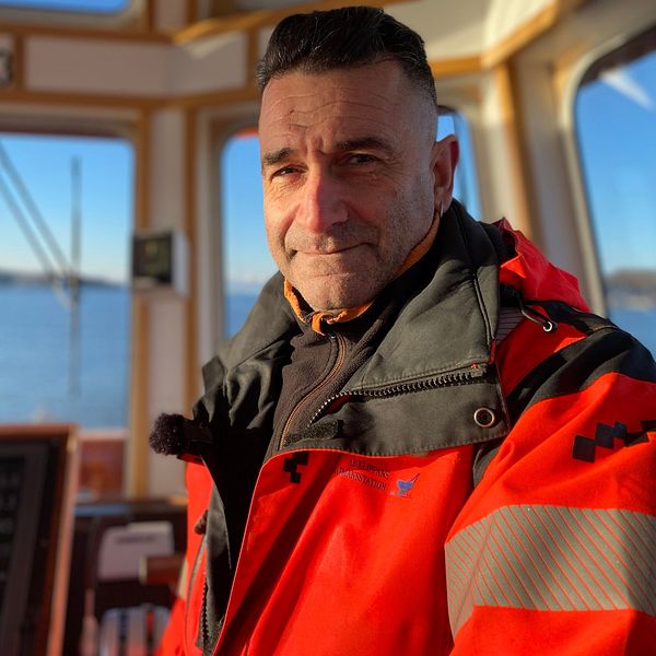 Man i orangea yrkeskläder står i fören på en båt och havet syns bakom honom.