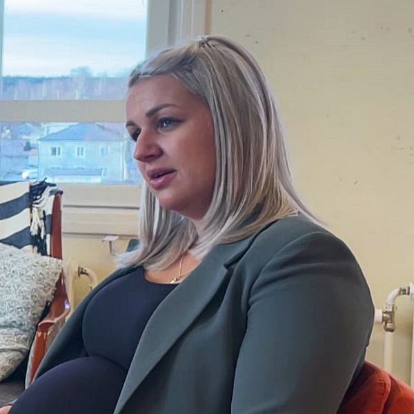 En gravid kvinna sitter på en stol med en grön kavaj och en svart topp, och hon ser ut över ett rum med fönster som ger utsikt mot en stadsmiljö. I bakgrunden finns en soffa med mönstrat tyg och en radiator. Rummet har en ljus och neutral inredning.