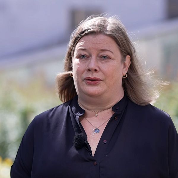 Person med långt blont hår står utomhus framför en grönskande bakgrund.