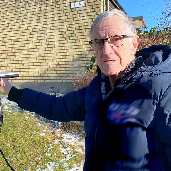 Person i vinterjacka pekar på elbilsladdare bredvid tegelbyggnad. Snö på marken.
