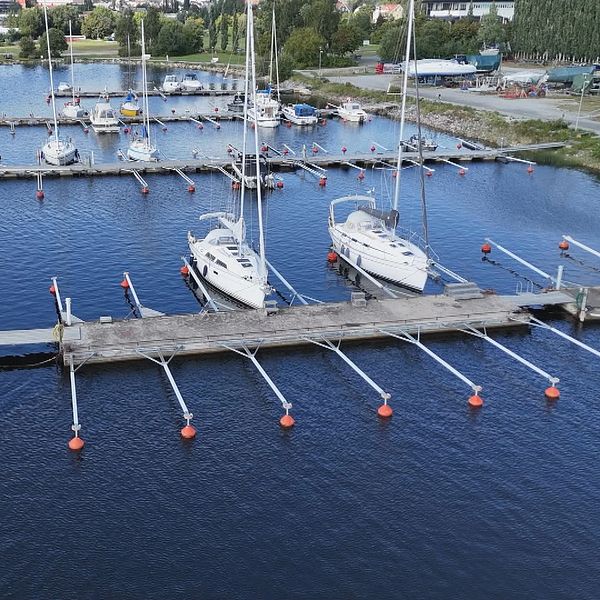 Flera segelbåtar är förtöjda vid en marin med flytbryggor, omgivna av stilla vatten. I förgrunden syns en blå båt vid kajen, medan flera andra båtar är fördelade längs med bryggan. I bakgrunden kan man skymta grönskande områden och byggnader.