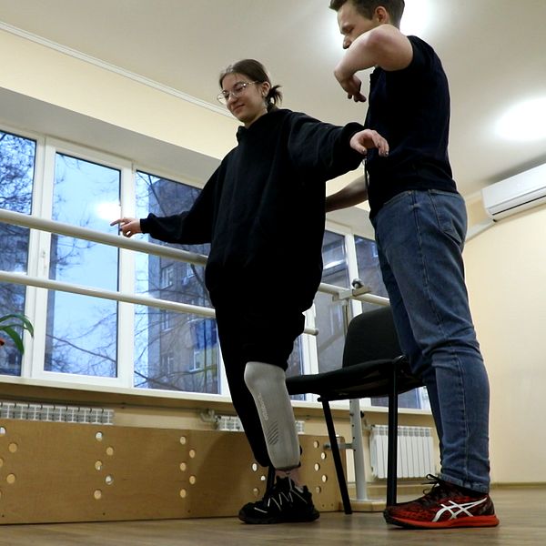 En person med protes står vid en balansstav med stöd av en annan person, i en rehabiliteringsmiljö.