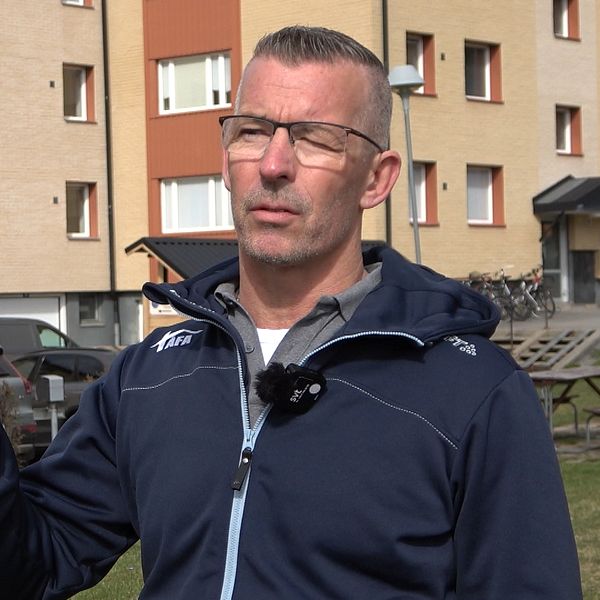 Man i blå jacka står utomhus framför ett flerfamiljshus, där AI styr inomhustemperaturen, och gör en gest med handen.
