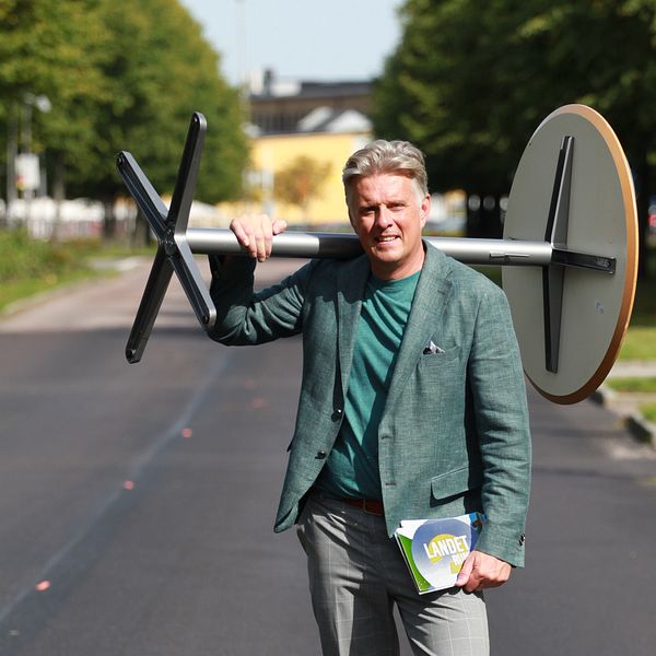 SVT:s programledare Henrik Kruusval står på en väg och har ett studiobord på axeln