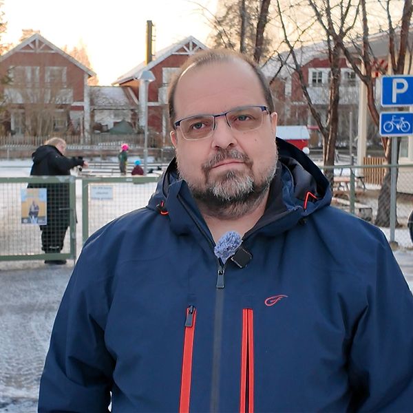 En person i mörk jacka står vid en grind som leder in till en snötäckt väg. I bakgrunden syns en parkering med en cykel och en person som går mot en annan som står vid en bänk. Området är omgiven av träd och hus, vilket skapar en vinterlig atmosfär. Solen går ner i horisonten, vilket ger en varm ton till bilden.