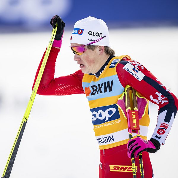 En längdskidåkare klädd i röd tävlingsdräkt med sponsorsloggor lutar sig mot en skidstav. Skidåkaren bär gula och röda handskar, en vit mössa med sponsormärken och en gul tävlingsväst märkt ”COOP FIS World Cup” samt ”ENGADIN”. Bakgrunden är snötäckt med en suddig banderoll med texten ”COOP”.