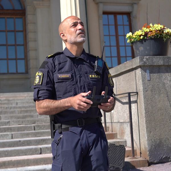 En uniformerad polis står framför en stentrappa med en handkontroll. Har blicken mot skyn.
