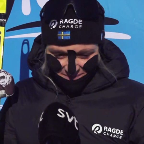En person i skidsportutrustning står framför en blå bakgrund med logotyper. Personen håller en skida och har på sig en svart jacka med sponsorlogotypen Ragde Charge. Det ser ut att vara en intervju efter en tävling.