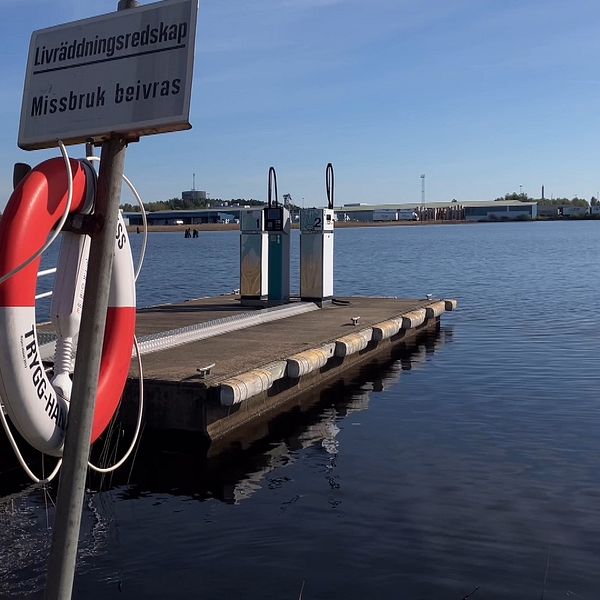 I förgrunden syns en röd och vit räddningsboj på en stolpe och i bakgrunden en sjömack med två pumpar på en brygga i Vänern.