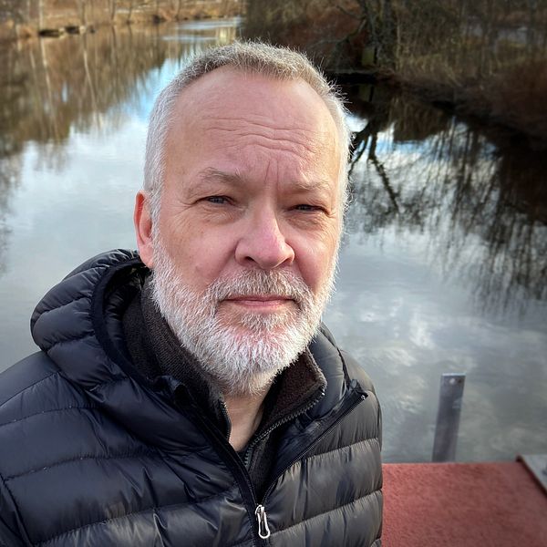 En person i svart jacka står på en bro med en spegelblanka sjö i bakgrunden, omgiven av träd och en lugn landskapsvy.