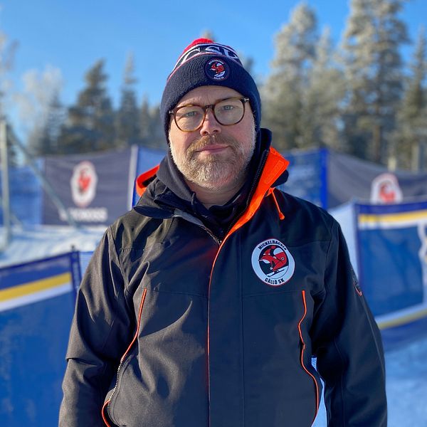 En man med lite skägg, glasögon, blå jacka och mössa, står utomhus på vintern framför skicrossbanan i Gällö