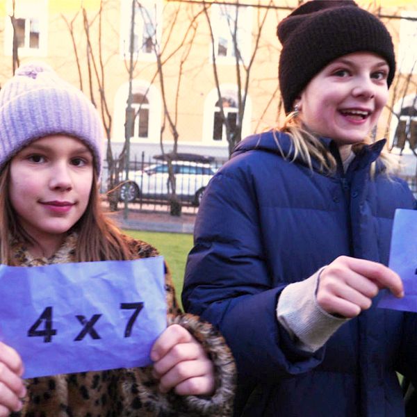 Barn håller upp färgglada kort med matematiska multiplikationsuppgifter utomhus. Tre av dem visar talen ”4 x 7”, ”6” och ”4 x 3”. Bakar och klädstilen tyder på en kall väderlek.