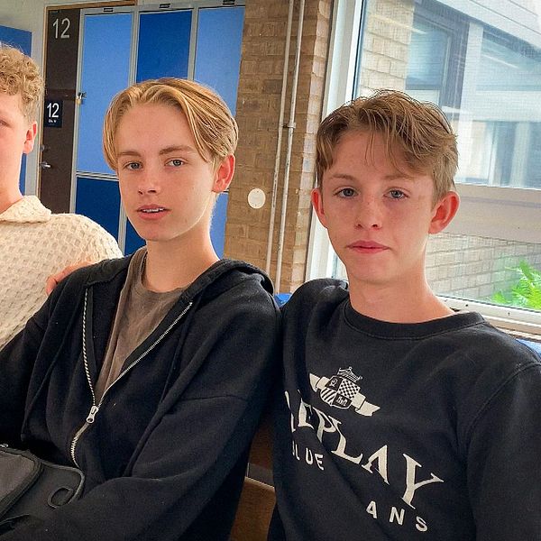 Tre killar sitter på en bänk i en skolkorridor.