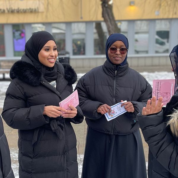 Fyra kvinnor i vinterkläder står utomhus och samtalar, några av dem håller papper i händerna. I bakgrunden syns en gul byggnad med fönster.