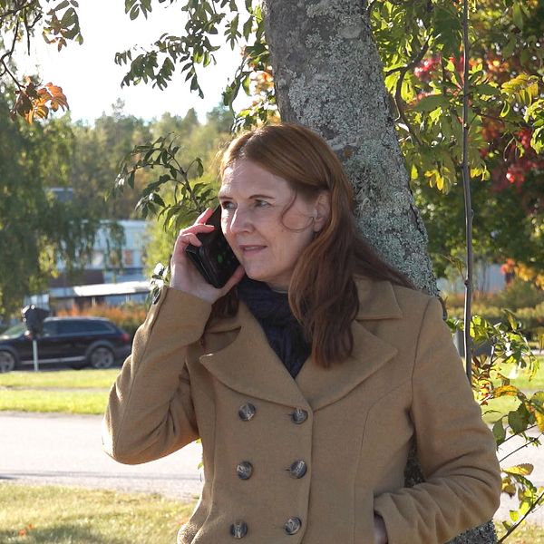 En person står lutad mot ett träd och pratar i telefon. Bakgrunden visar en park med grönska och fler människor som promenerar. Det är en solig dag med klar himmel och höstlöv i olika färger.
