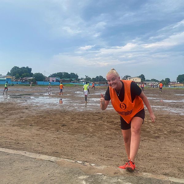 Idrottsvolontär Clara Engstrand gör tummen upp framför en lerig fotbollsplan i Kongo-Brazzaville
