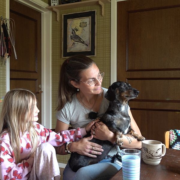 I ett 70-tals kök vid borde sitter en liten tjej och hennes mamma som har en tax-hund i knät