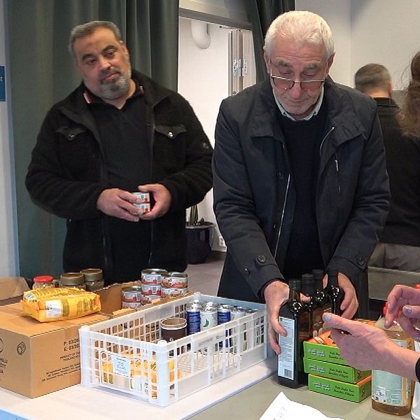 Två män i svarta jackor packar upp matvaror på ett bord. En kvinna med telefon köper en vara.
