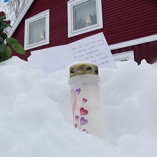Ett minnesmärke i snön utanför ett rött hus. Märket består av ett blockljus dekorerat med hjärtan, en bukett röda rosor och ett handskrivet brev.