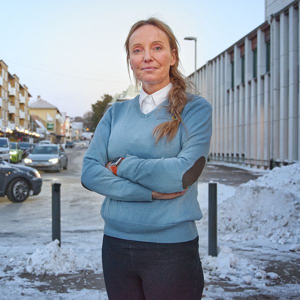 En person står med armarna i kors på en gata i en stad, omgiven av snö. I bakgrunden syns flerbostadshus och flera bilar parkerade längs gatan.