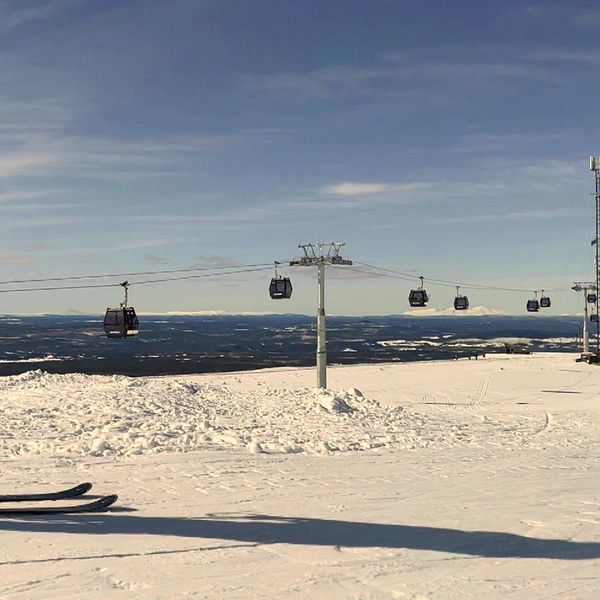 En skidåkare i grön jacka står på en snötäckt backe och blickar mot en fjärran utsikt, medan skidliftar, gondoler, svävar över landskapet i bakgrunden.