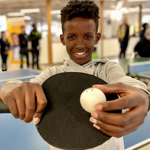 Ung person håller ett pingisracket och boll, med personer i bakgrunden.
