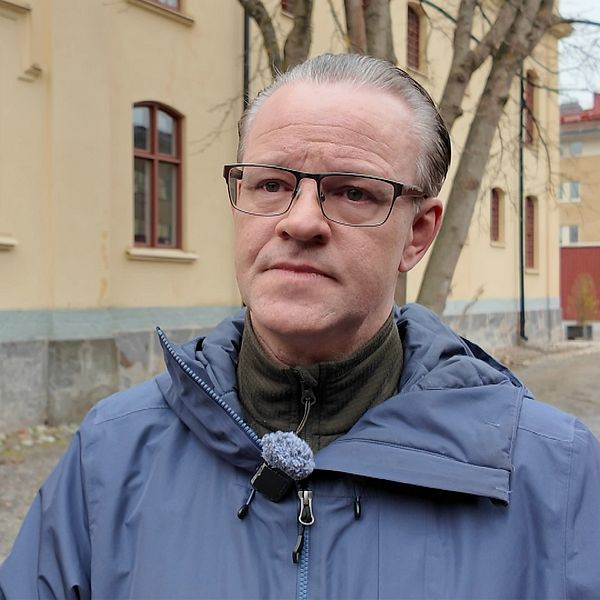 Person utomhus framför gulputsad byggnad med röda fönster och träd i bakgrunden.