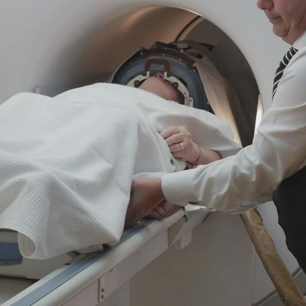 En patient ligger på en bår som förs in i en MRI-maskin, med två vårdpersonal som hjälper till. En person är klädd i en skjorta med lång ärm och en annan i en vit skjorta och mörk väst. Patienten är täckt med en filtar och har en huvudskyddsanordning på sig.