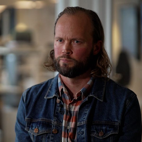 En person sitter framåtlutad i en kontorsmiljö, iförd en jeansjacka och en rutig skjorta. I bakgrunden syns oskarpa kontorsdetaljer och datorer.