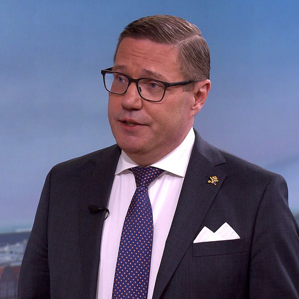 Man i kostym och slips intervjuas i tv-studio med stadsutsikt i bakgrunden.