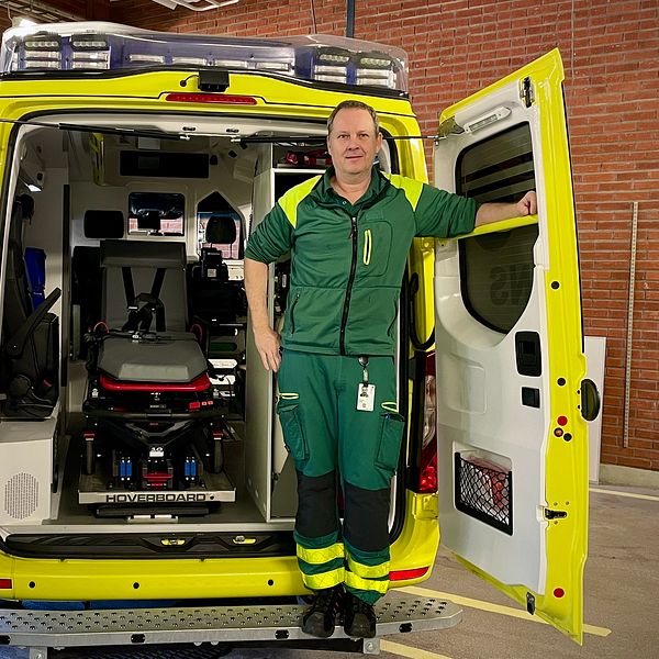En ambulans med öppen bakdörr visar insidan som har en bår och medicinsk utrustning. En person i gröna arbetskläder står vid ambulansens öppna dörrar.