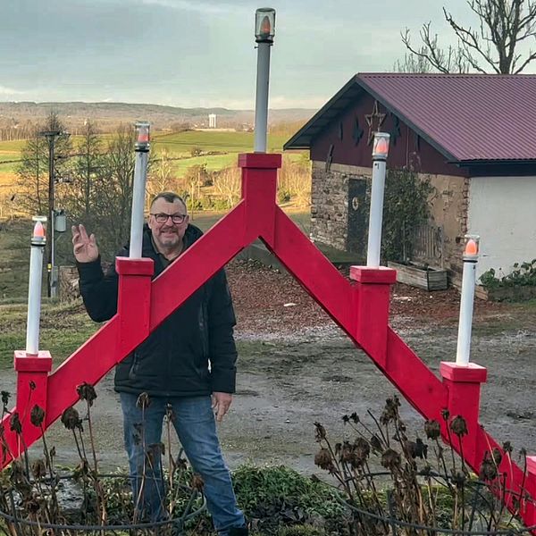 En person står framför en stor, röd dekoration i form av en stor adventsljusstake. I bakgrunden syns en landskapsvy med grönskande ängar och träd, samt en byggnad med röd tak. Vädret ser klart ut med en ljus himmel.