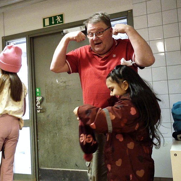 En skola, där barn förbereder sig för att gå ut i en kapphall. Ett barn tar på sig en jacka medan en annan klär på sig i bakgrunden. Några personliga tillhörigheter finns på hyllorna längs väggen. En vuxen person står i dörröppningen och blir uppmärksammad av barnen.