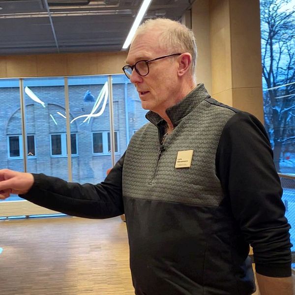 En man pekar mot en av flera skåp i en modern byggnad med stora fönster, som ger en utsikt mot utsidan.