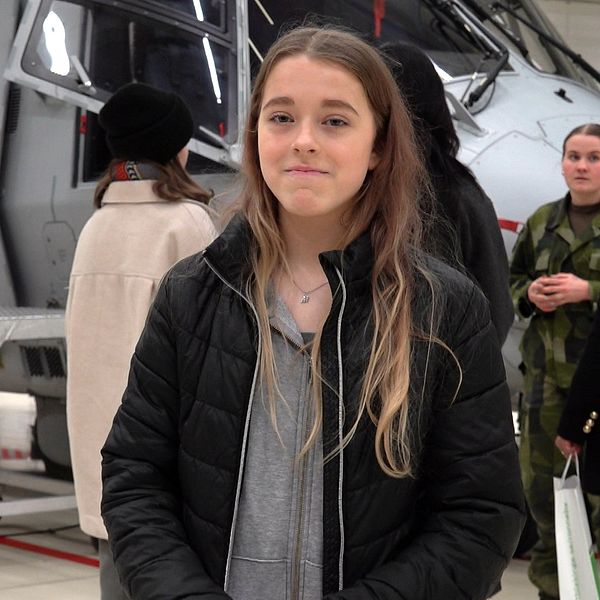 En grupp personer står framför en grå militärhelikopter inomhus. Helikoptern har öppen dörr och en stege som leder upp till den. En person bär en camouflagedräkt.