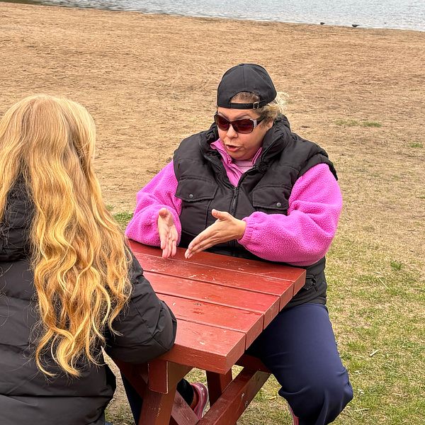 Två personer samtalar vid ett rött picknickbord utomhus nära vatten.