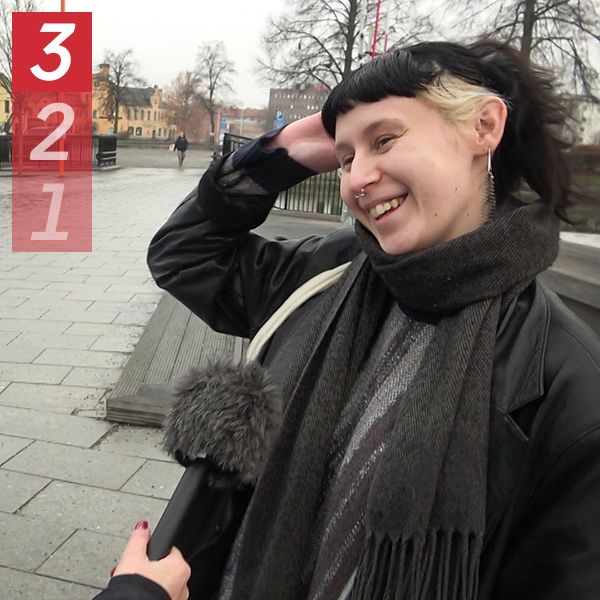 En person med en mörk jacka och en halsduk sträcker sig efter sitt hår medan en annan person, som håller i en mikrofon, ställer frågor. Scenen utspelar sig utomhus på en grå och molnig dag, med träd och byggnader i bakgrunden.